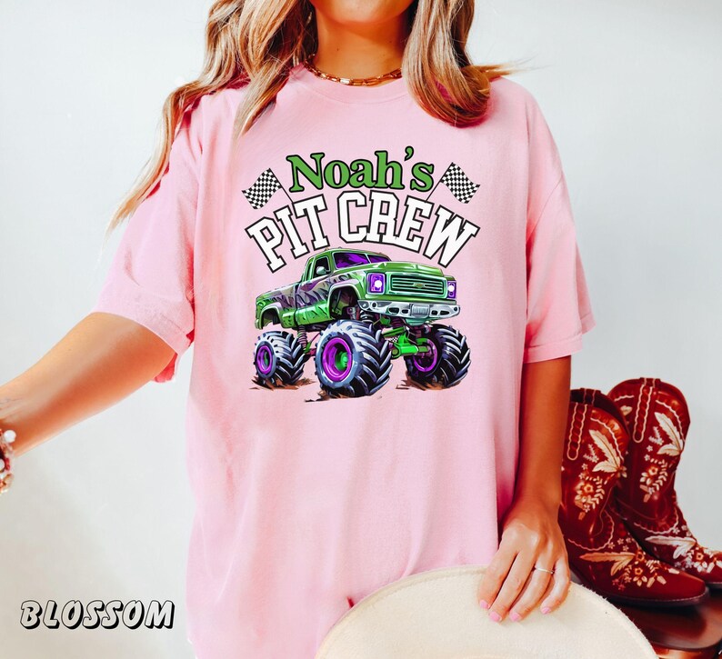 Pode incluir: Camiseta rosa claro com um desenho de um monster truck verde com rodas roxas e o texto "Noah's Pit Crew" em verde e branco. A camiseta tamb&eacute;m tem duas bandeiras quadriculadas.
