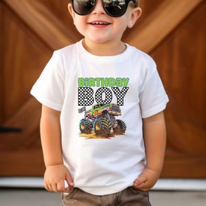 May include: White t-shirt with a colorful monster truck graphic and the words "BIRTHDAY BOY" in green and black checkered font. The monster truck has large blue tires and is driving on dirt.