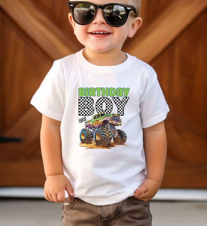 May include: White t-shirt with a colorful monster truck graphic and the words "BIRTHDAY BOY" in green and black checkered font. The monster truck has large blue tires and is driving on dirt.