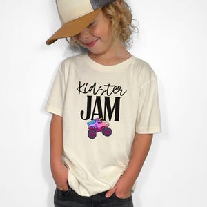 May include: Cream-colored t-shirt with the words "Kidster JAM" in black script and block letters. Below the text is a colorful monster truck with a unicorn design. The shirt is worn by a person wearing a baseball cap.