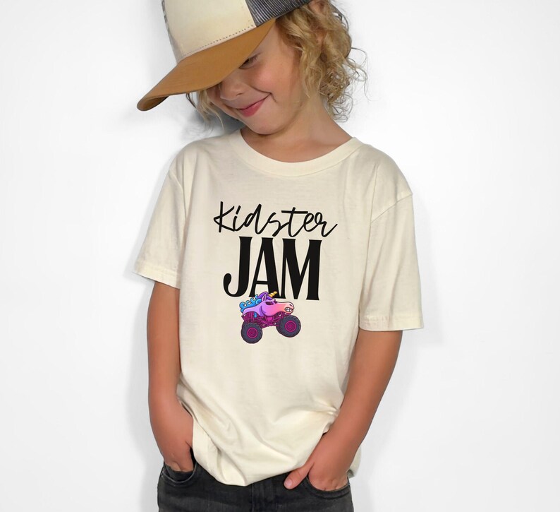 May include: Cream-colored t-shirt with the words "Kidster JAM" in black script and block letters. Below the text is a colorful monster truck with a unicorn design. The shirt is worn by a person wearing a baseball cap.