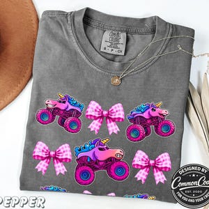 Pictured is a dark gray comfort colors t-shirt with three rows of alternating sparkle pink smash monster trucks and pink checkered bows