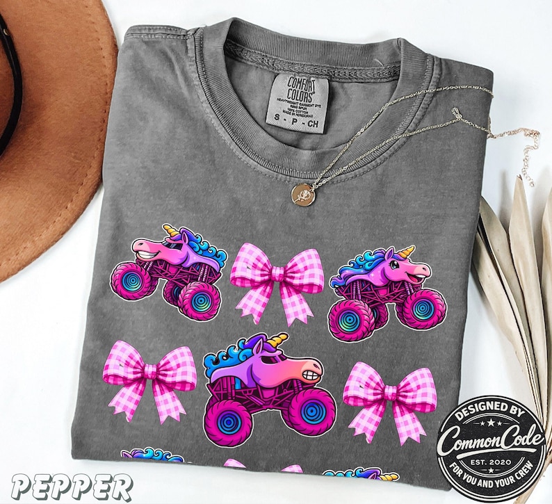 Pictured is a dark gray comfort colors t-shirt with three rows of alternating sparkle pink smash monster trucks and pink checkered bows
