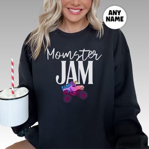 May include: Black sweatshirt with the words "Momster JAM" in white script and block letters. A colorful graphic of a unicorn monster truck is below the text. A white circle with the words "ANY NAME" is in the upper right corner.