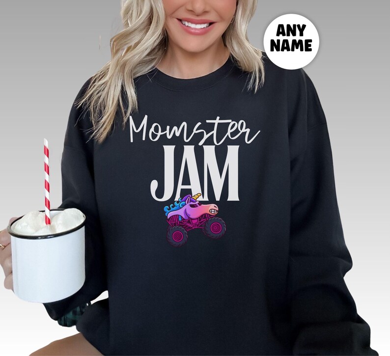 May include: Black sweatshirt with the words "Momster JAM" in white script and block letters. A colorful graphic of a unicorn monster truck is below the text. A white circle with the words "ANY NAME" is in the upper right corner.