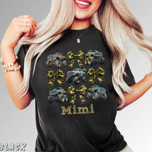 May include: Black t-shirt with a design of monster trucks and camouflage bows. The word "Mimi" is printed in gold lettering. The shirt is a comfortable, casual style.