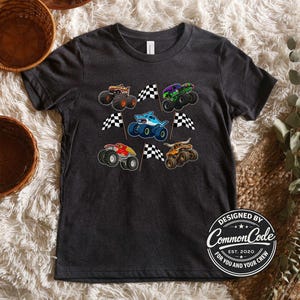 May include: Dark gray t-shirt featuring colorful monster truck graphics and checkered flags. The shirt includes a shark-themed truck and a logo that reads "Designed by Common Code Est. 2020 For You and Your Crew."