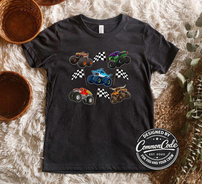May include: Dark gray t-shirt featuring colorful monster truck graphics and checkered flags. The shirt includes a shark-themed truck and a logo that reads "Designed by Common Code Est. 2020 For You and Your Crew."