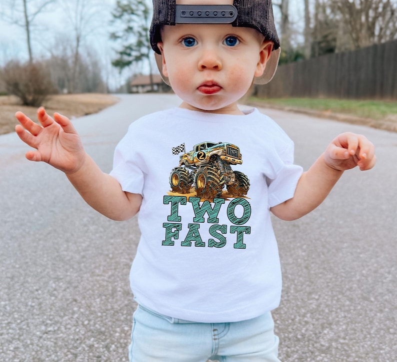 Pictured is a young boy on his second birthday wearing a white t_shirt which says TWO FAST in blue sketch letters along with a large monster truck graphic