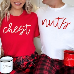 May include: A couple wearing matching red and white Christmas themed shirts. The woman's shirt says "chest" in white script and the man's shirt says "nuts" in red script. They are both wearing red and black plaid pajamas.