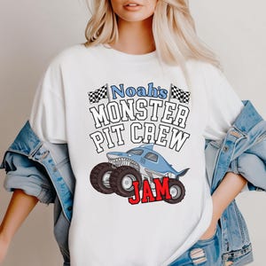 Puede incluir: Camiseta blanca con un dise&ntilde;o de cami&oacute;n monstruo azul con forma de tibur&oacute;n, ruedas negras y el texto "Noah's Monster Pit Crew Jam". El dise&ntilde;o incluye banderas a cuadros y una fuente l&uacute;dica.