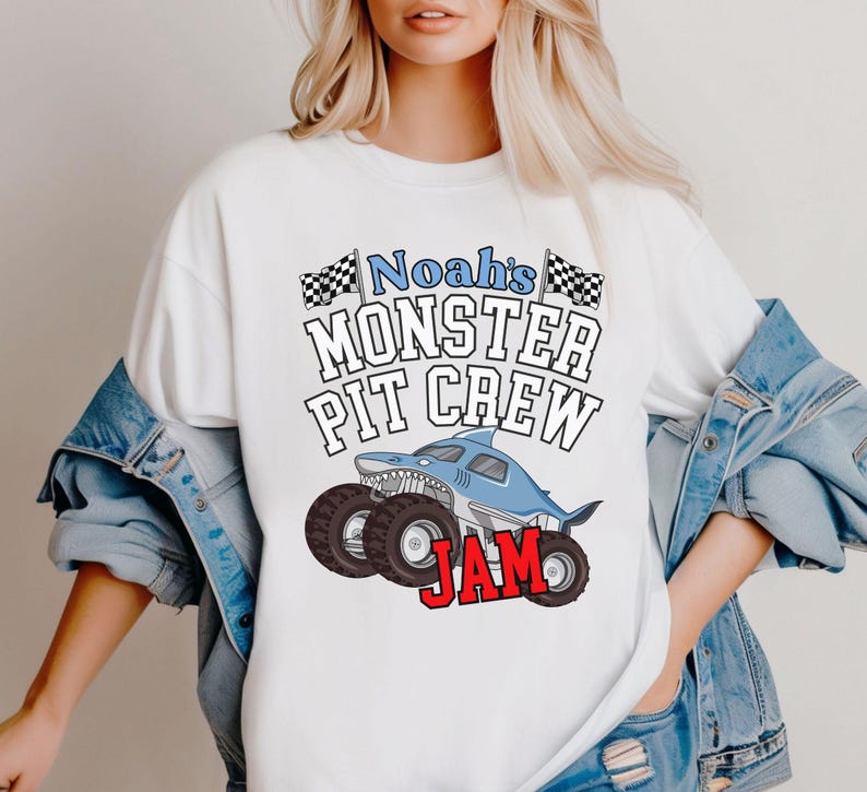 Puede incluir: Camiseta blanca con un dise&ntilde;o de cami&oacute;n monstruo azul con forma de tibur&oacute;n, ruedas negras y el texto "Noah's Monster Pit Crew Jam". El dise&ntilde;o incluye banderas a cuadros y una fuente l&uacute;dica.