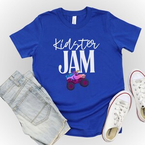 May include: A royal blue t-shirt with the words "Kidster JAM" in white script. A cartoon monster truck with a unicorn head is featured below the text. The shirt is paired with denim shorts and white canvas sneakers.