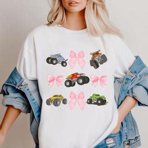 K&ouml;nnte beinhalten: Wei&szlig;es T-Shirt mit farbenfrohen Illustrationen von Monstertrucks und rosa Schleifen. Die Monstertrucks sind in Blau, Orange, Rot, Gelb und Gr&uuml;n gehalten. Die Schleifen sind rosa.