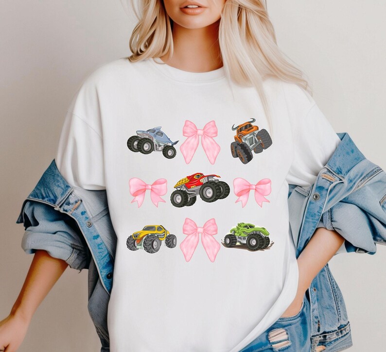 K&ouml;nnte beinhalten: Wei&szlig;es T-Shirt mit farbenfrohen Illustrationen von Monstertrucks und rosa Schleifen. Die Monstertrucks sind in Blau, Orange, Rot, Gelb und Gr&uuml;n gehalten. Die Schleifen sind rosa.
