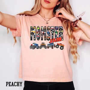 K&ouml;nnte beinhalten: Ein pfirsichfarbenes T-Shirt mit dem Aufdruck "MOMSTER JAM" in Schwarz. Die Buchstaben bestehen aus Bildern von Monstertrucks.