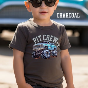 May include: Charcoal gray t-shirt featuring a blue monster truck graphic with the words "PIT CREW" above it. The monster truck has large tires and a checkered flag. The word "CHARCOAL" is printed in white.