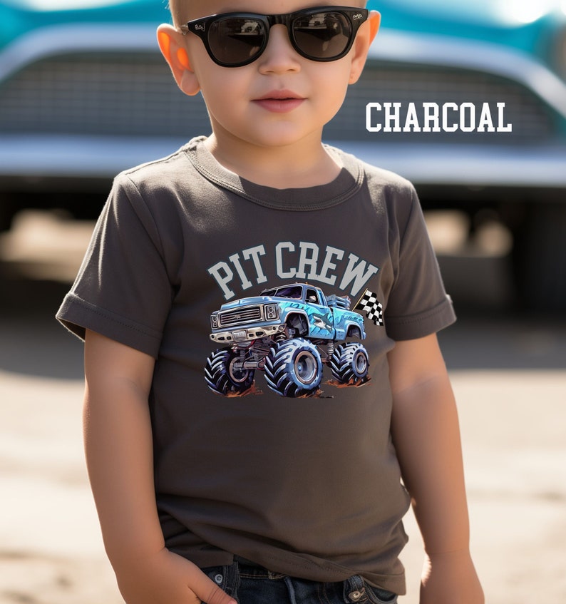 May include: Charcoal gray t-shirt featuring a blue monster truck graphic with the words "PIT CREW" above it. The monster truck has large tires and a checkered flag. The word "CHARCOAL" is printed in white.