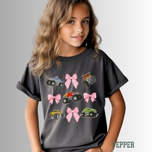 K&ouml;nnte beinhalten: Ein anthrazitfarbenes T-Shirt mit gestickten Monstertrucks in verschiedenen Farben, darunter Blau, Orange, Rot, Gelb und Gr&uuml;n. Jeder Truck ist mit einer rosa Schleife versehen. Das Shirt ist ein bequemer, l&auml;ssiger Stil.