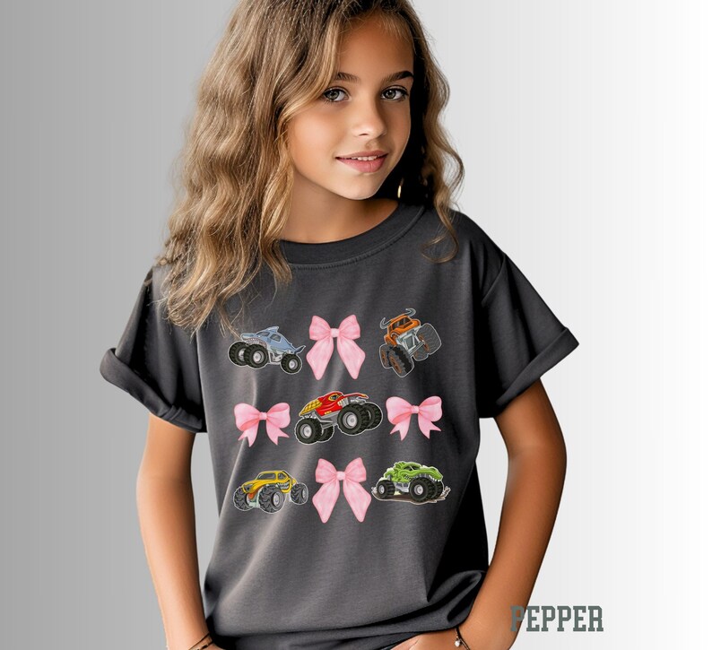 K&ouml;nnte beinhalten: Ein anthrazitfarbenes T-Shirt mit gestickten Monstertrucks in verschiedenen Farben, darunter Blau, Orange, Rot, Gelb und Gr&uuml;n. Jeder Truck ist mit einer rosa Schleife versehen. Das Shirt ist ein bequemer, l&auml;ssiger Stil.
