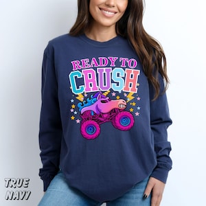 May include: Navy blue long-sleeve sweatshirt with the text "READY TO CRUSH" above a cartoon unicorn monster truck. The graphic features pink, purple, and blue colors. The sweatshirt is made of a soft material.