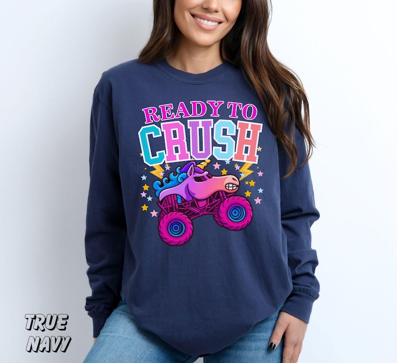 May include: Navy blue long-sleeve sweatshirt with the text "READY TO CRUSH" above a cartoon unicorn monster truck. The graphic features pink, purple, and blue colors. The sweatshirt is made of a soft material.