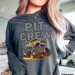 May include: Dark gray long-sleeve shirt with the words "PIT CREW" in a checkered pattern. A colorful monster truck graphic is below the text. The truck is green, purple, and yellow with large blue tires.