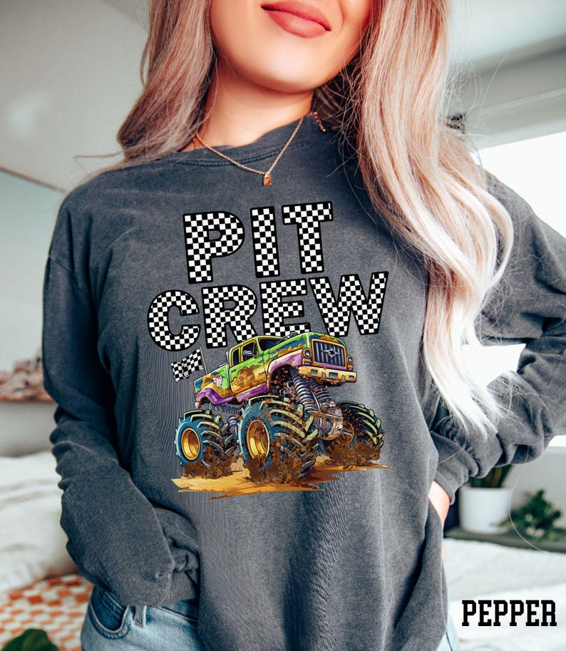 May include: Dark gray long-sleeve shirt with the words "PIT CREW" in a checkered pattern. A colorful monster truck graphic is below the text. The truck is green, purple, and yellow with large blue tires.