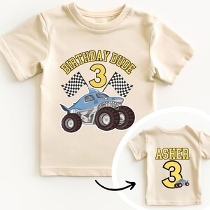 May include: Cream-colored t-shirt with a shark-themed monster truck graphic and the text "BIRTHDAY DUDE 3". The back of the shirt has the name "ASHER" and the number "3".