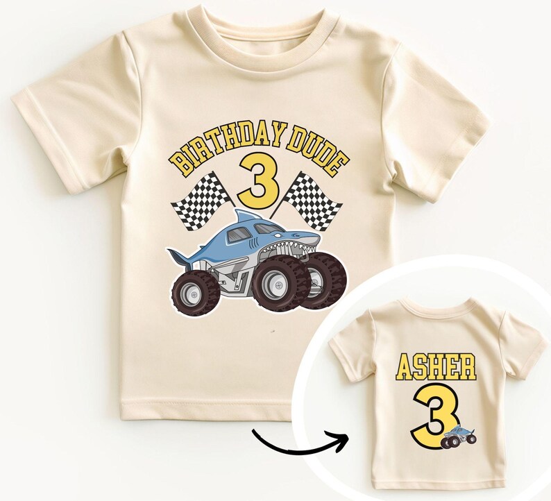 May include: Cream-colored t-shirt with a shark-themed monster truck graphic and the text "BIRTHDAY DUDE 3". The back of the shirt has the name "ASHER" and the number "3".