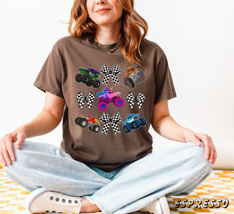 May include: A brown t-shirt featuring colorful monster truck graphics and checkered bow accents. The trucks are in various colors, including green, orange, pink, and blue. The shirt is worn by a person.