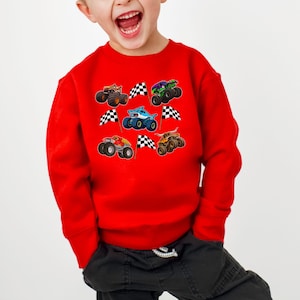May include: A red sweatshirt featuring colorful monster truck graphics and checkered flags. The design includes various monster trucks in shades of brown, green, blue, and orange. The sweatshirt is a long-sleeved crew neck.