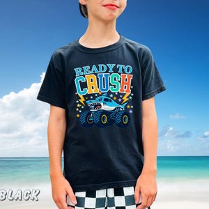 May include: Black t-shirt with a graphic of a shark monster truck and the text "READY TO CRUSH". The design includes lightning bolts and stars. The shirt is worn by a person against a blue sky background.
