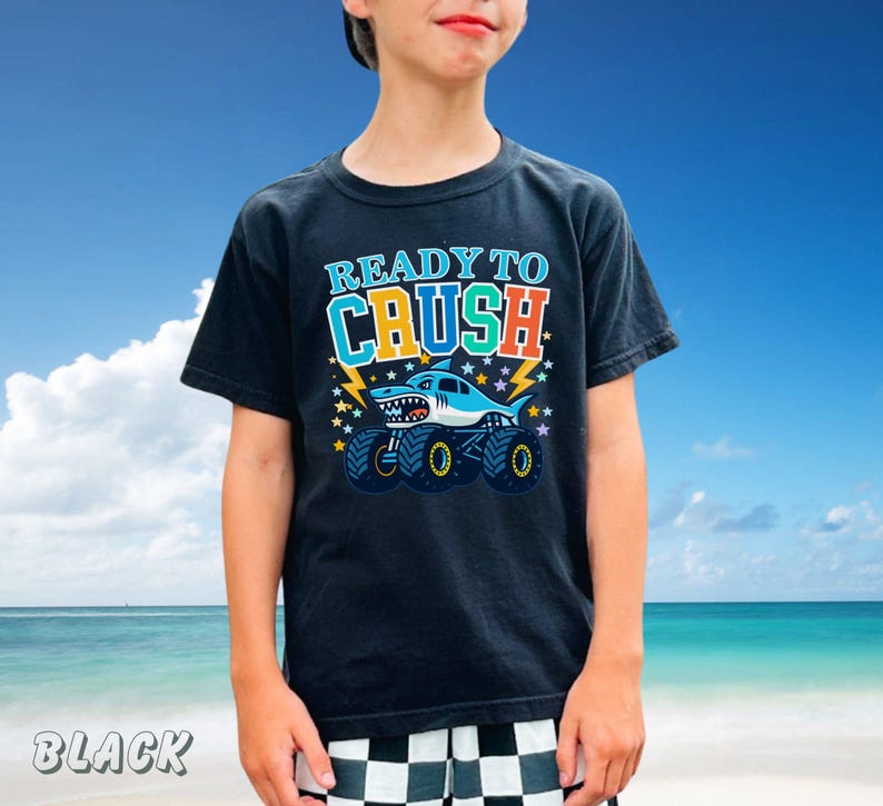 May include: Black t-shirt with a graphic of a shark monster truck and the text "READY TO CRUSH". The design includes lightning bolts and stars. The shirt is worn by a person against a blue sky background.