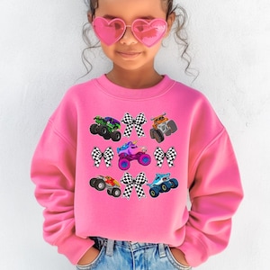 Op de afbeelding: Een felroze sweatshirt met kleurrijke monstertruckafbeeldingen en geblokte strikken. Het ontwerp bevat groene, paarse, oranje, rode en blauwe monstertrucks. De sweatshirt is gemaakt van een zacht materiaal.
