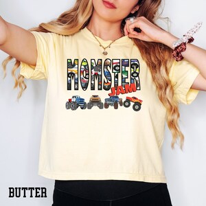 K&ouml;nnte beinhalten: Ein gelbes T-Shirt mit dem Text "Momster Jam" in schwarzen Buchstaben. Die Buchstaben bestehen aus Bildern von Monstertrucks. Am unteren Rand des Shirts befinden sich f&uuml;nf Monstertrucks in einer Reihe.