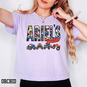 K&ouml;nnte beinhalten: Ein lavendelfarbenes T-Shirt mit dem Text "Ariel's Jam" in schwarzen Buchstaben. Die Buchstaben bestehen aus Bildern von Monstertrucks.