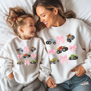 May include: White sweatshirts with colorful monster truck graphics and pink bow accents. The sweatshirts are worn by a mother and child, creating a matching set. The monster trucks are in various colors, including blue, orange, red, yellow, and green.
