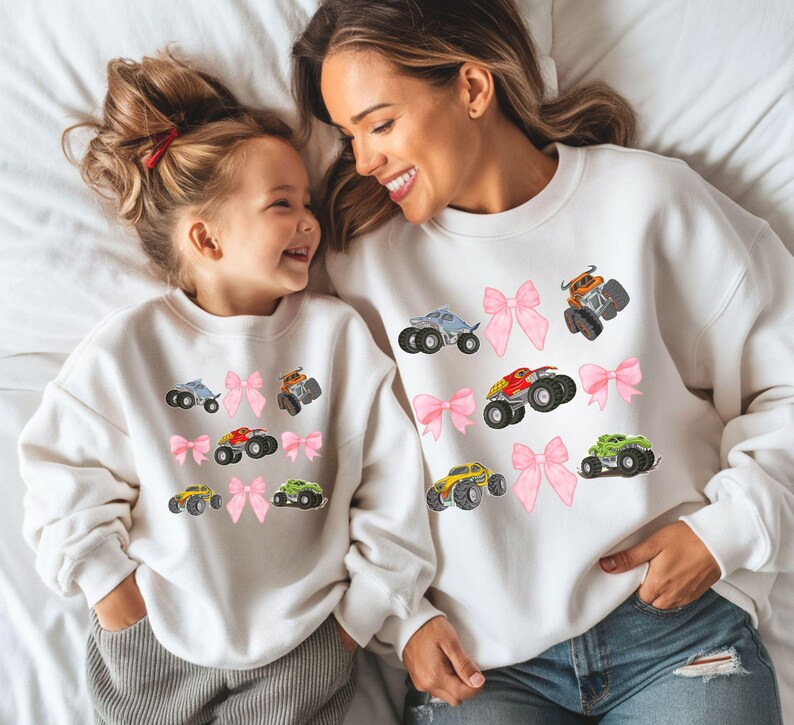 May include: White sweatshirts with colorful monster truck graphics and pink bow accents. The sweatshirts are worn by a mother and child, creating a matching set. The monster trucks are in various colors, including blue, orange, red, yellow, and green.