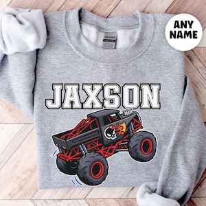 Puede incluir: Una sudadera gris con el nombre "JAXSON" en blanco y negro sobre un gráfico de un camión monstruo rojo y negro. El camión tiene un diseño de calavera y llamas. La sudadera tiene cuello redondo y mangas largas.