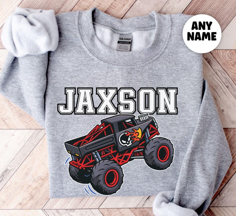 a grey sweatshirt that says "Jaxson" in black and white varsity style letters, below is a red and black monster truck with the punisher on it and a fire flames. Bone Shaker themed truck.  There is a white circle on the page that says "ANY NAME" - custom names , with infant, toddler, kid and adult sizes.