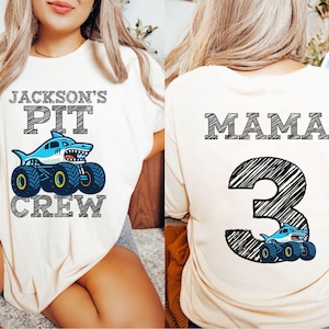 pictured is a woman wearing a cream-colored t-shirt with the text "JACKSON'S PIT CREW" above a shark-themed monster truck graphic. The back of the shirt features the word "MAMA" and the number "3" with a shark truck graphic.