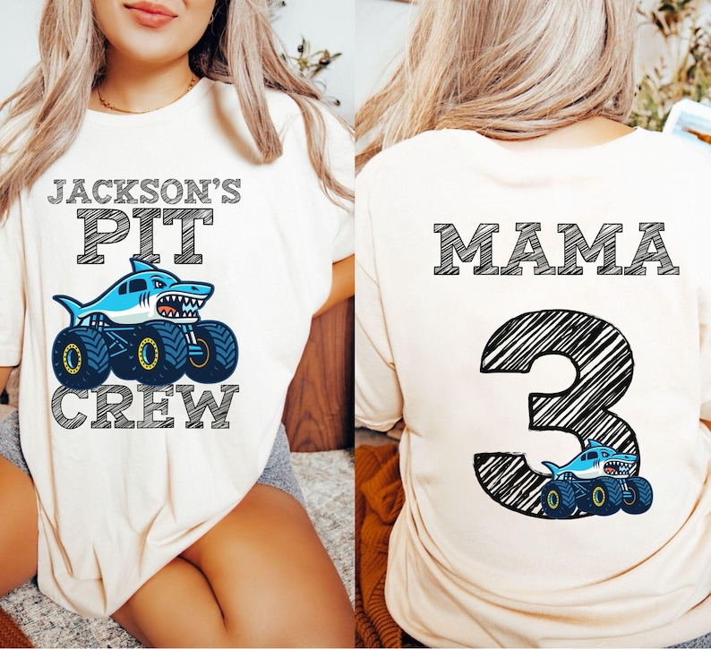 pictured is a woman wearing a cream-colored t-shirt with the text "JACKSON'S PIT CREW" above a shark-themed monster truck graphic. The back of the shirt features the word "MAMA" and the number "3" with a shark truck graphic.