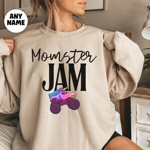 May include: Beige long-sleeve sweatshirt with the text "Momster JAM" in black script and block letters. A colorful monster truck graphic with a unicorn horn is below the text. A white circle with the words "ANY NAME" is in the upper left corner.