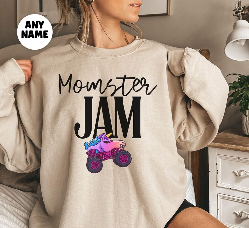 May include: Beige long-sleeve sweatshirt with the text "Momster JAM" in black script and block letters. A colorful monster truck graphic with a unicorn horn is below the text. A white circle with the words "ANY NAME" is in the upper left corner.