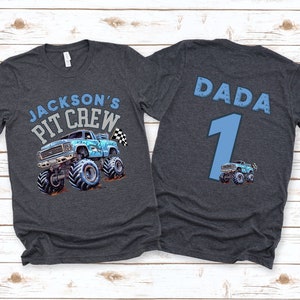 May include: Dark grey t-shirt featuring a blue monster truck graphic and the text "JACKSON'S PIT CREW" on the front. The back of the shirt has the word "DADA", the number "1", and a small monster truck graphic.