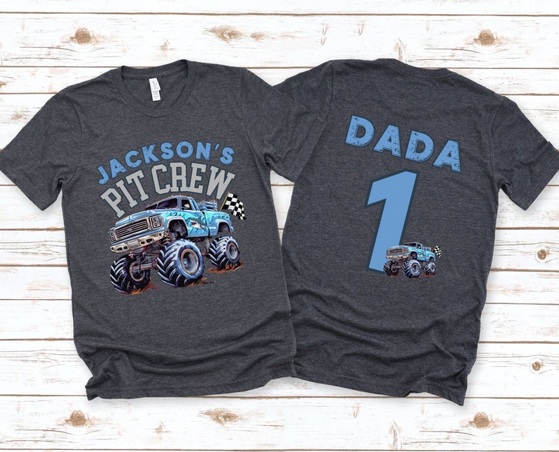 May include: Dark grey t-shirt featuring a blue monster truck graphic and the text "JACKSON'S PIT CREW" on the front. The back of the shirt has the word "DADA", the number "1", and a small monster truck graphic.