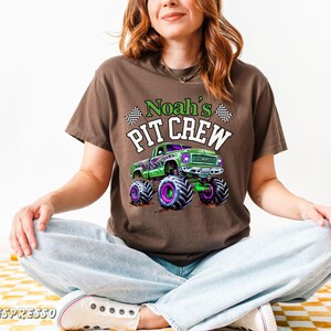 Pode incluir: Camiseta marrom com um design de monster truck verde e detalhes roxos e pretos. O texto "Noah's PIT CREW" &eacute; impresso acima do caminh&atilde;o. A camiseta &eacute; usada por uma pessoa sentada de pernas cruzadas.