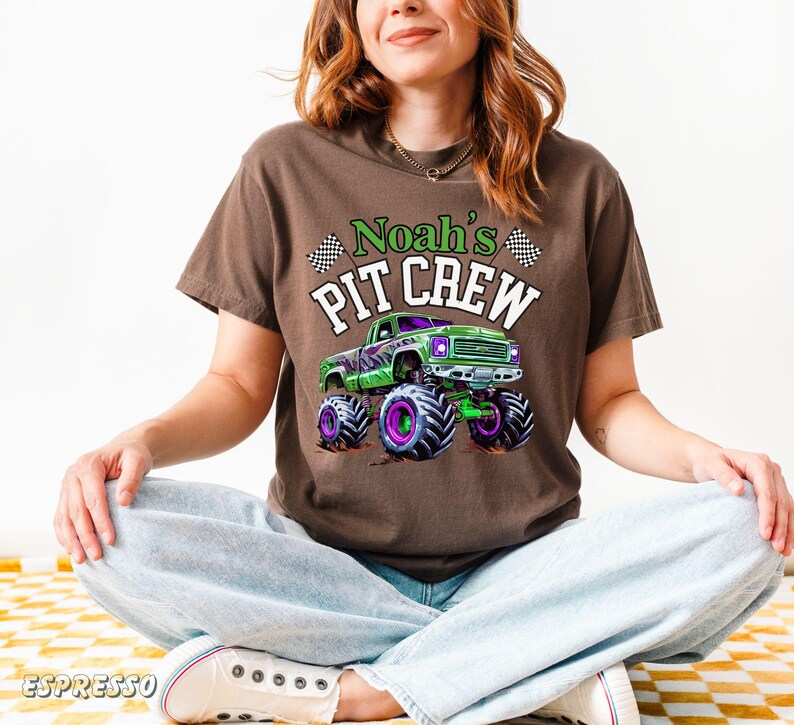 Pode incluir: Camiseta marrom com um design de monster truck verde e detalhes roxos e pretos. O texto "Noah's PIT CREW" &eacute; impresso acima do caminh&atilde;o. A camiseta &eacute; usada por uma pessoa sentada de pernas cruzadas.
