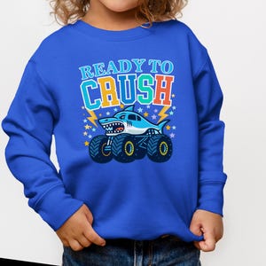 Pu&ograve; includere: Una felpa blu con una grafica di un monster truck a forma di squalo e la scritta "READY TO CRUSH". Lo squalo &egrave; azzurro e bianco. Il testo &egrave; in arancione, verde e blu.